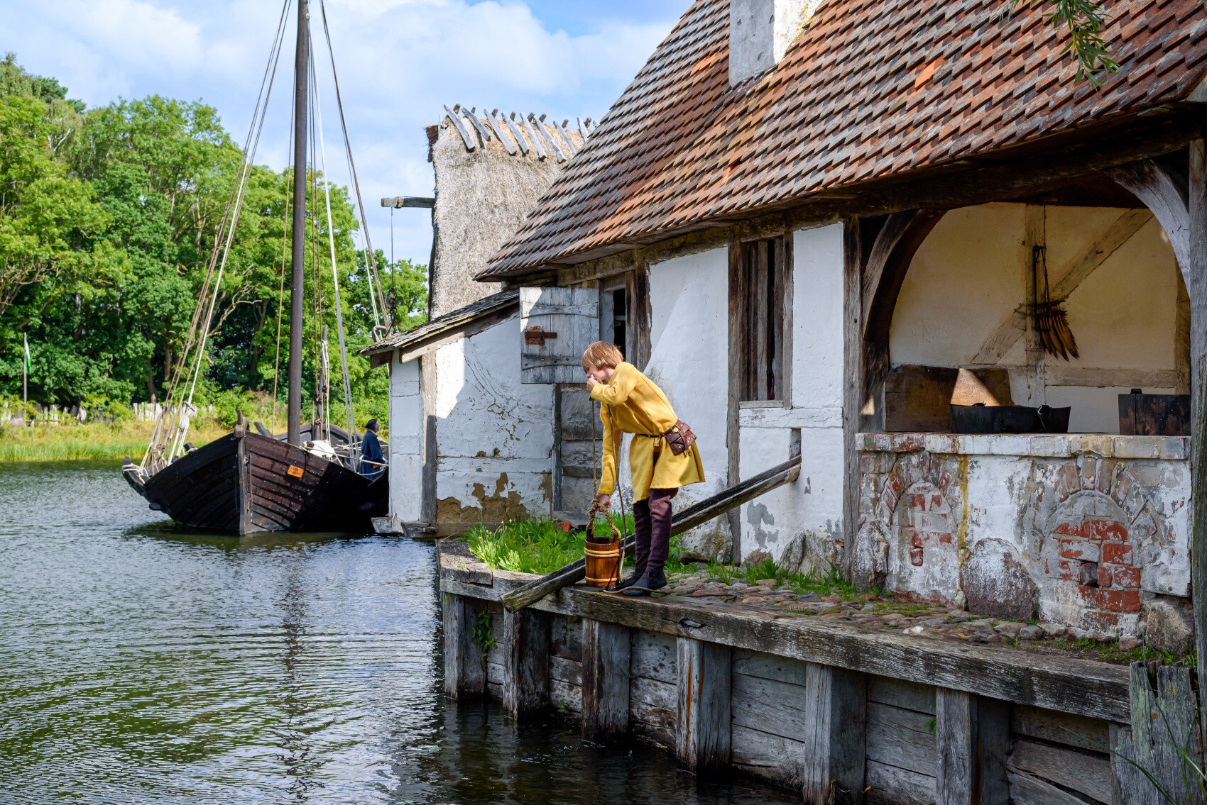 Ein Kind in mittelalterlicher Kleidung seilt einen Holzeimer hinab in einen Fluss in einem Freilichtmuseum Ein Kind in mittelalterlicher Kleidung seilt einen Holzeimer hinab in einen Fluss in einem Freilichtmuseum