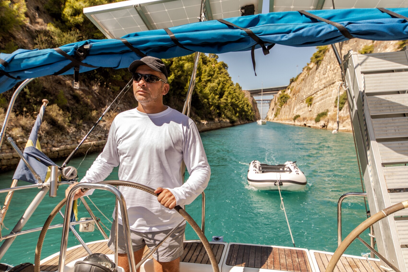 Ein Mann am Steuer eines Segelbootes auf einer künstlichen Wasserstraße, gefolgt von weiteren Booten Ein Mann am Steuer eines Segelbootes auf einer künstlichen Wasserstraße, gefolgt von weiteren Booten