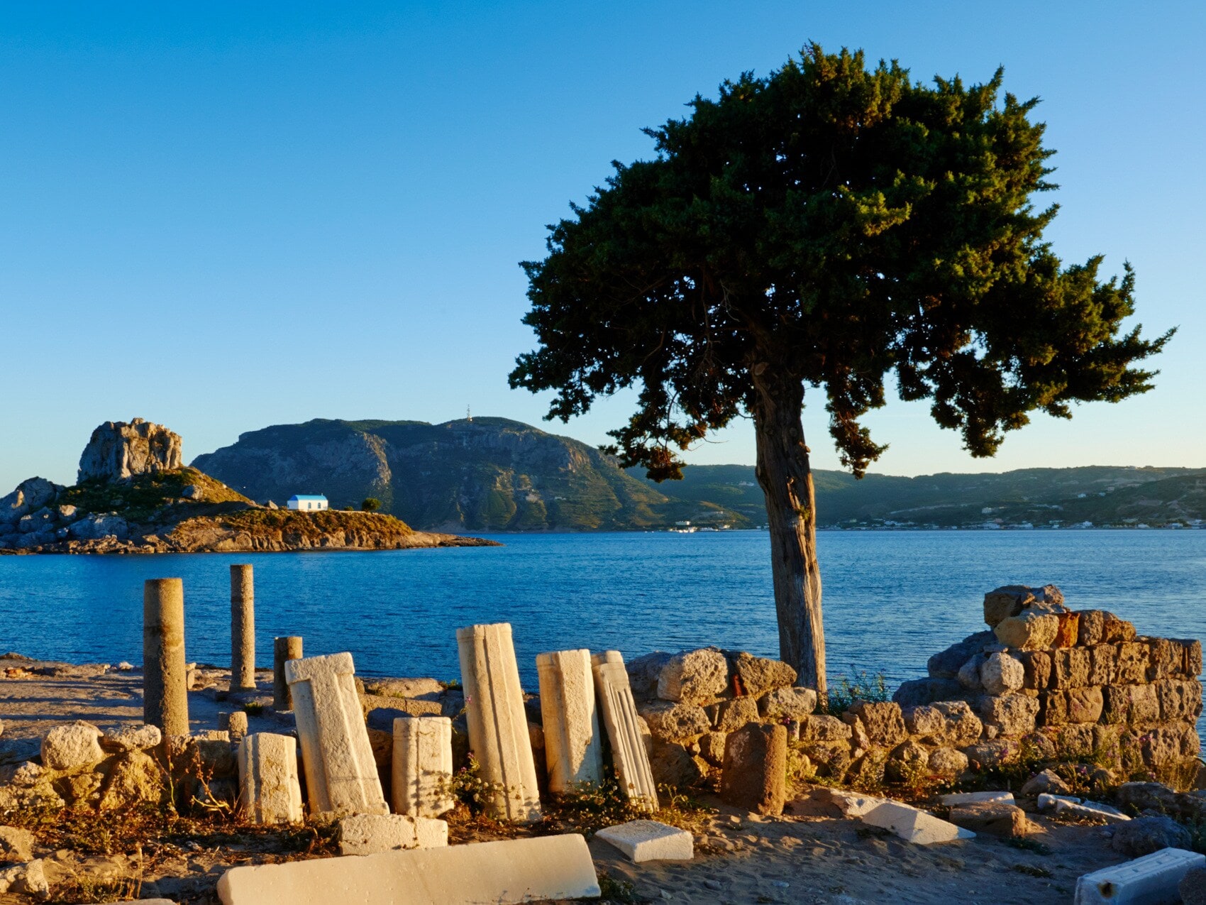 Ruinenstätte mit Überresten von Säulen in einer Meeresbucht mit Blick auf eine vorgelagerte Felsinsel bei Sonnenuntergang Ruinenstätte mit Überresten von Säulen in einer Meeresbucht mit Blick auf eine vorgelagerte Felsinsel bei Sonnenuntergang