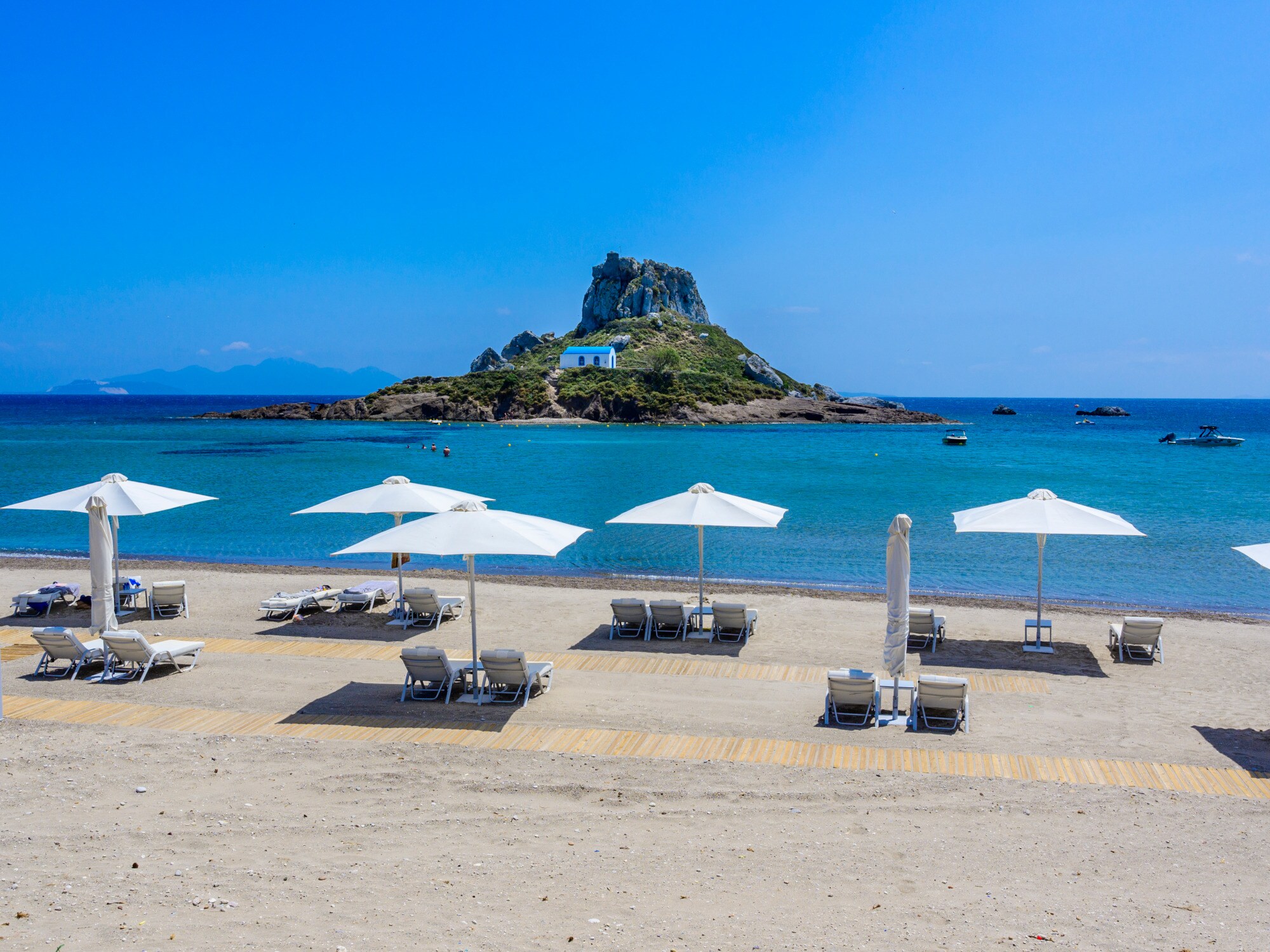 Aufgereihte Liegen mit Sonnenschirmen an einem Sandstrand gegenüber einer kleinen Felsinsel mit orthodoxer Kirche Aufgereihte Liegen mit Sonnenschirmen an einem Sandstrand gegenüber einer kleinen Felsinsel mit orthodoxer Kirche
