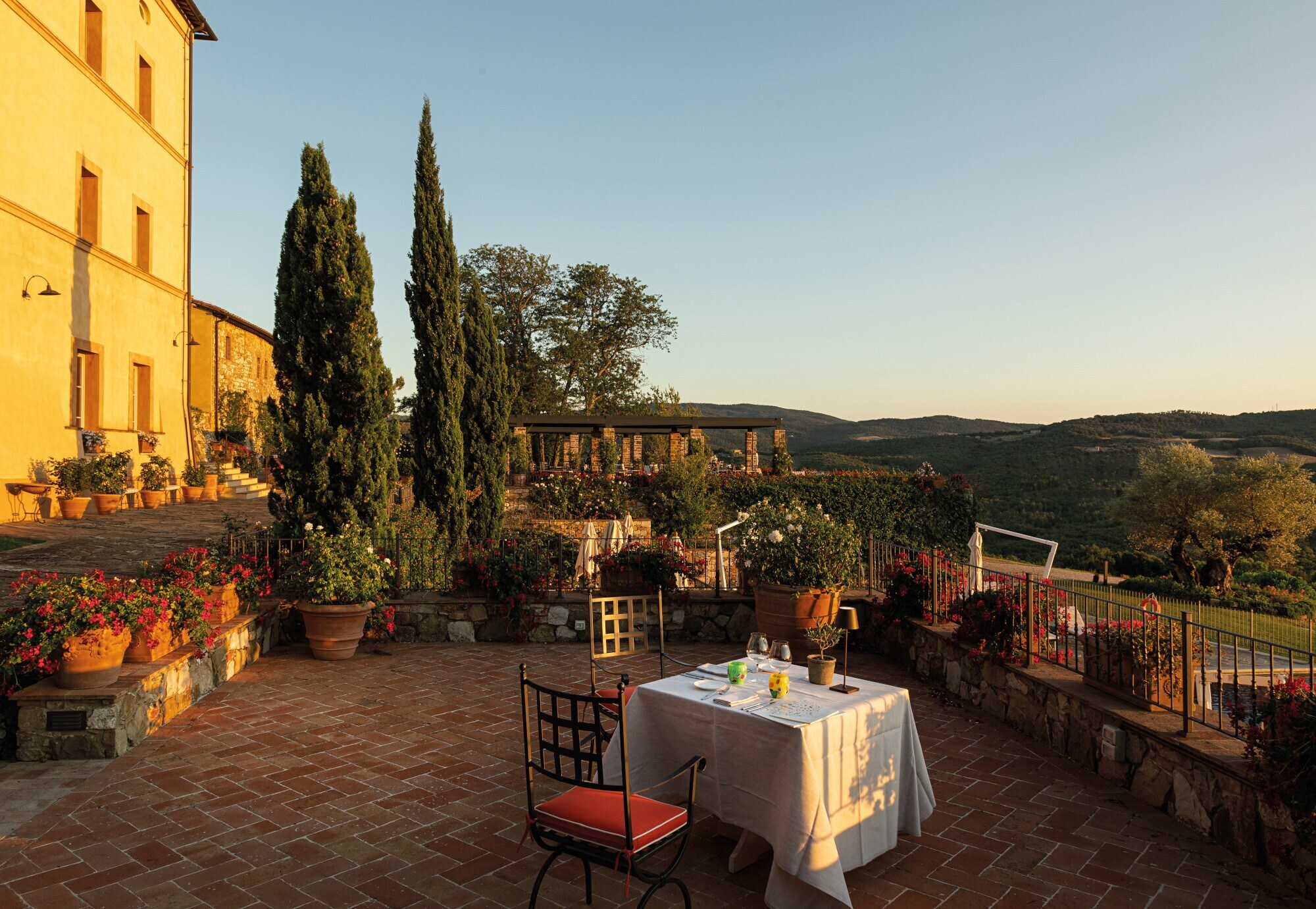 Ein gedeckter Tisch auf einer großen Terrasse einer Villa im toskanischen Baustil mit Blick in eine grüne Hügellandschaft bei Sonnenuntergang
