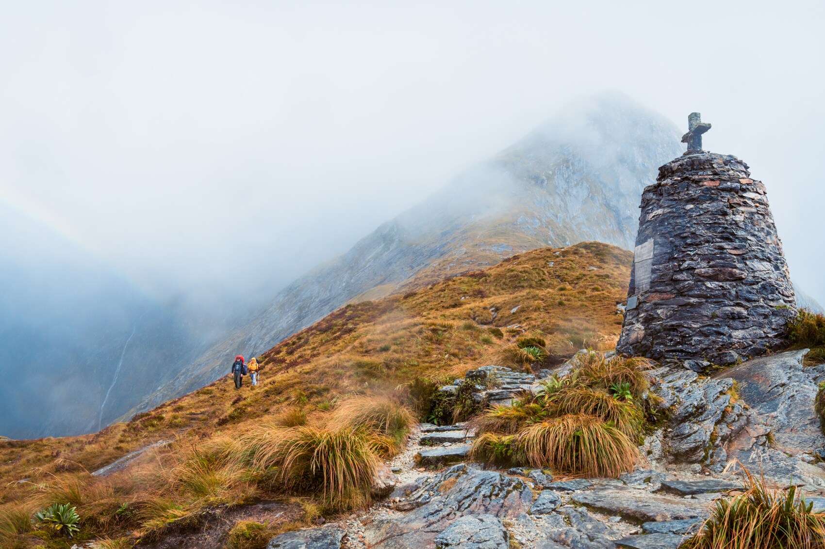 Zwei Wandernde auf dem Milford Track zwischen von Nebel umgebenen Felsen und Graslandschaft