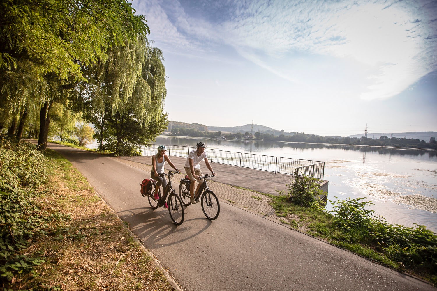 Ein Paar in Sommerkleidung fährt auf Fahrrädern auf einem Radweg entlang eines Flusses Ein Paar in Sommerkleidung fährt auf Fahrrädern auf einem Radweg entlang eines Flusses