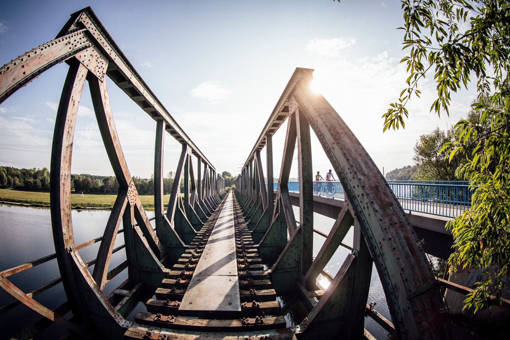 Zwei Radfahrer fahren auf einer Brücke neben einer alten Eisenbahnbrücke über einen Fluss