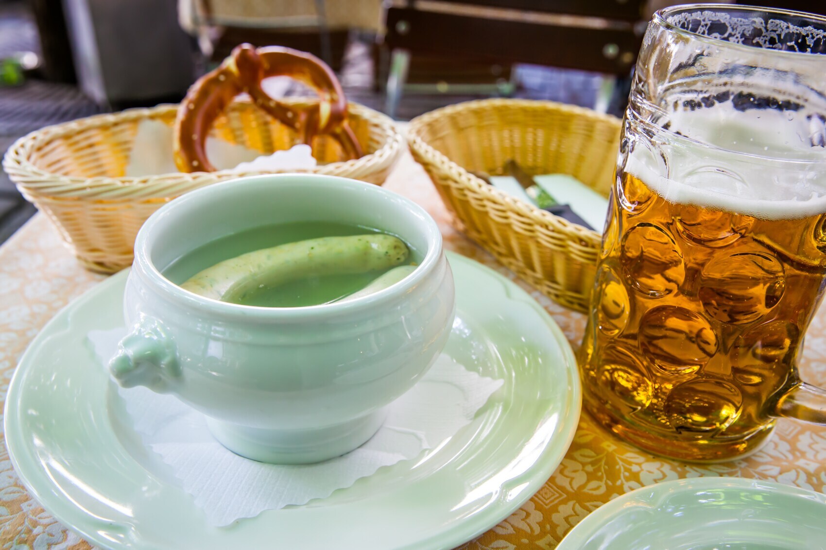 Tisch mit einem Suppenteller, gefüllt mit zwei Weißwürsten, dahinter ein Korb mit einer Brezel, daneben ein Glas Bier Tisch mit einem Suppenteller, gefüllt mit zwei Weißwürsten, dahinter ein Korb mit einer Brezel, daneben ein Glas Bier