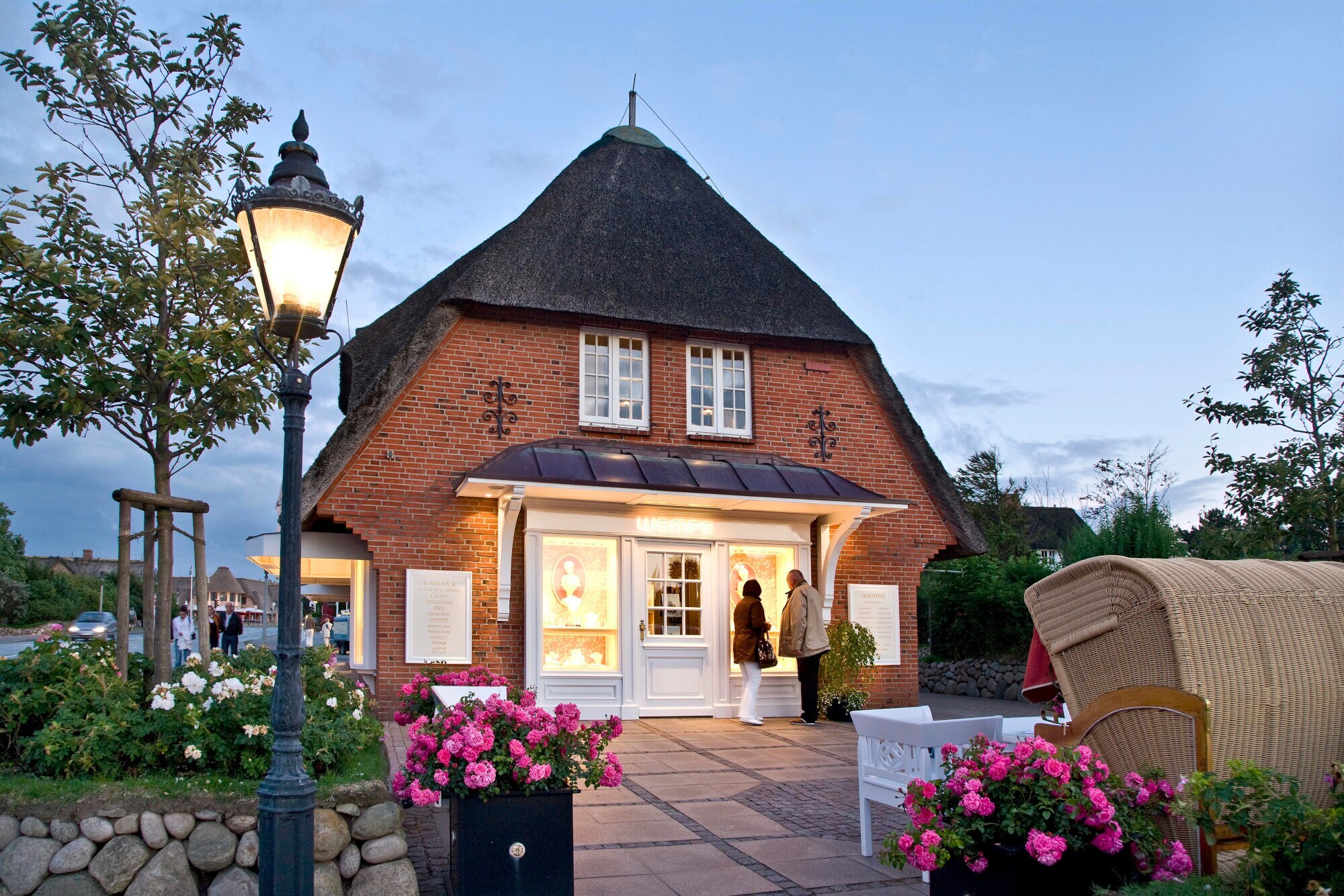 Zwei Personen stehen vor einem beleuchteten Schaufenster einer Luxusboutique in einem reetgedeckten Haus Zwei Personen stehen vor einem beleuchteten Schaufenster einer Luxusboutique in einem reetgedeckten Haus