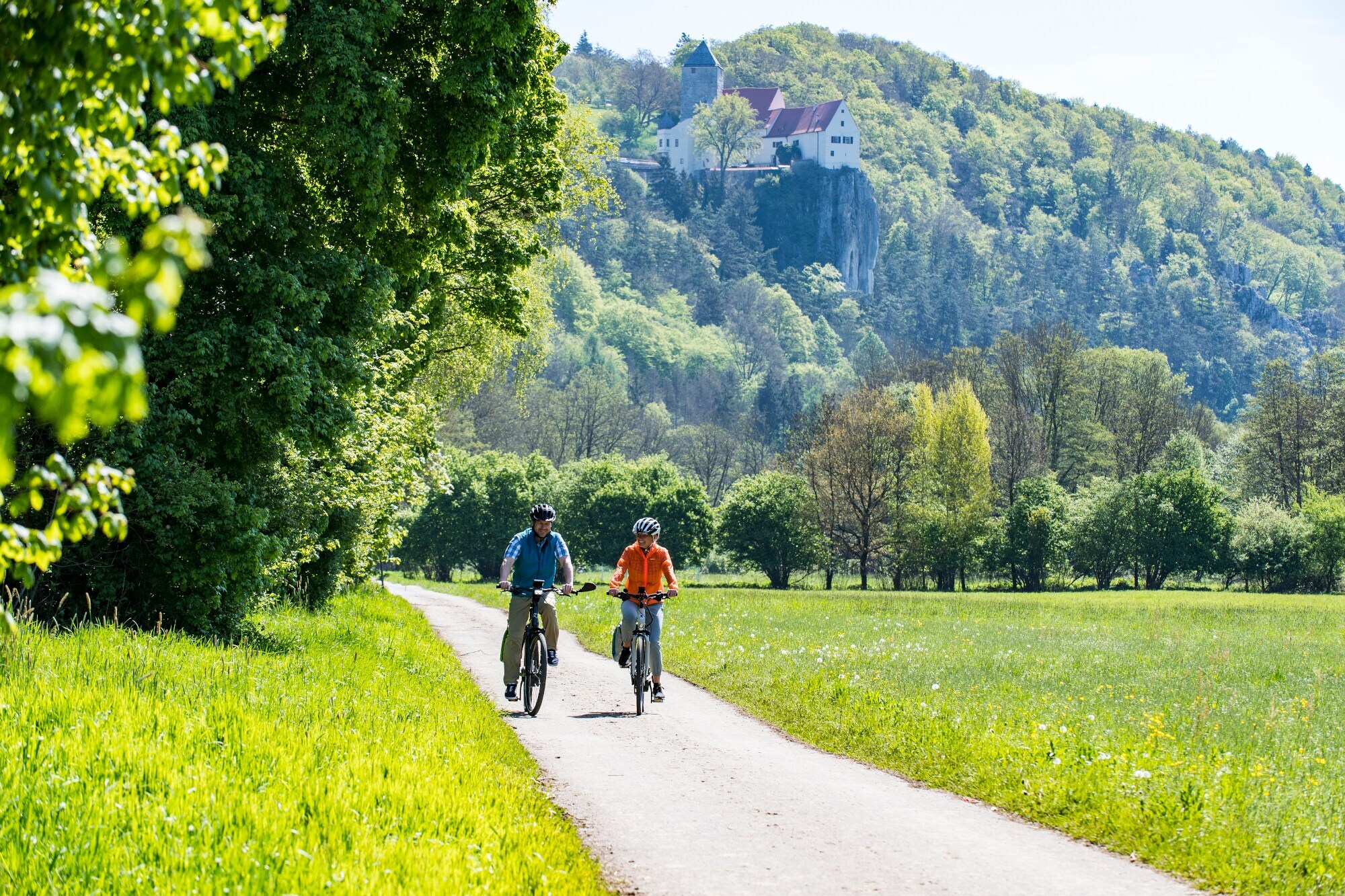 Zwei Personen auf Fahrrädern fahren auf einem Radweg durch ein grünes Tal.