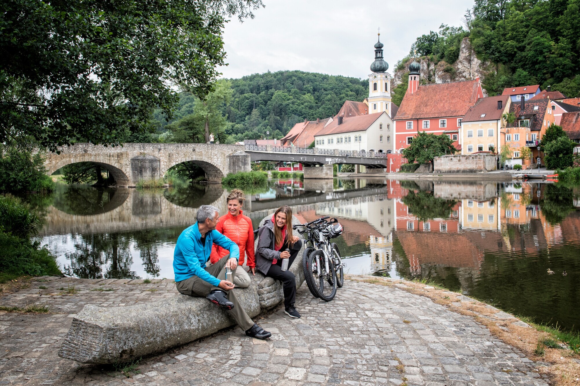 Drei Personen mit Fahrrädern sitzen auf einem Stein am Ufer eines Flusses vor dem Panorama einer mittelalterlichen Kleinstadt.