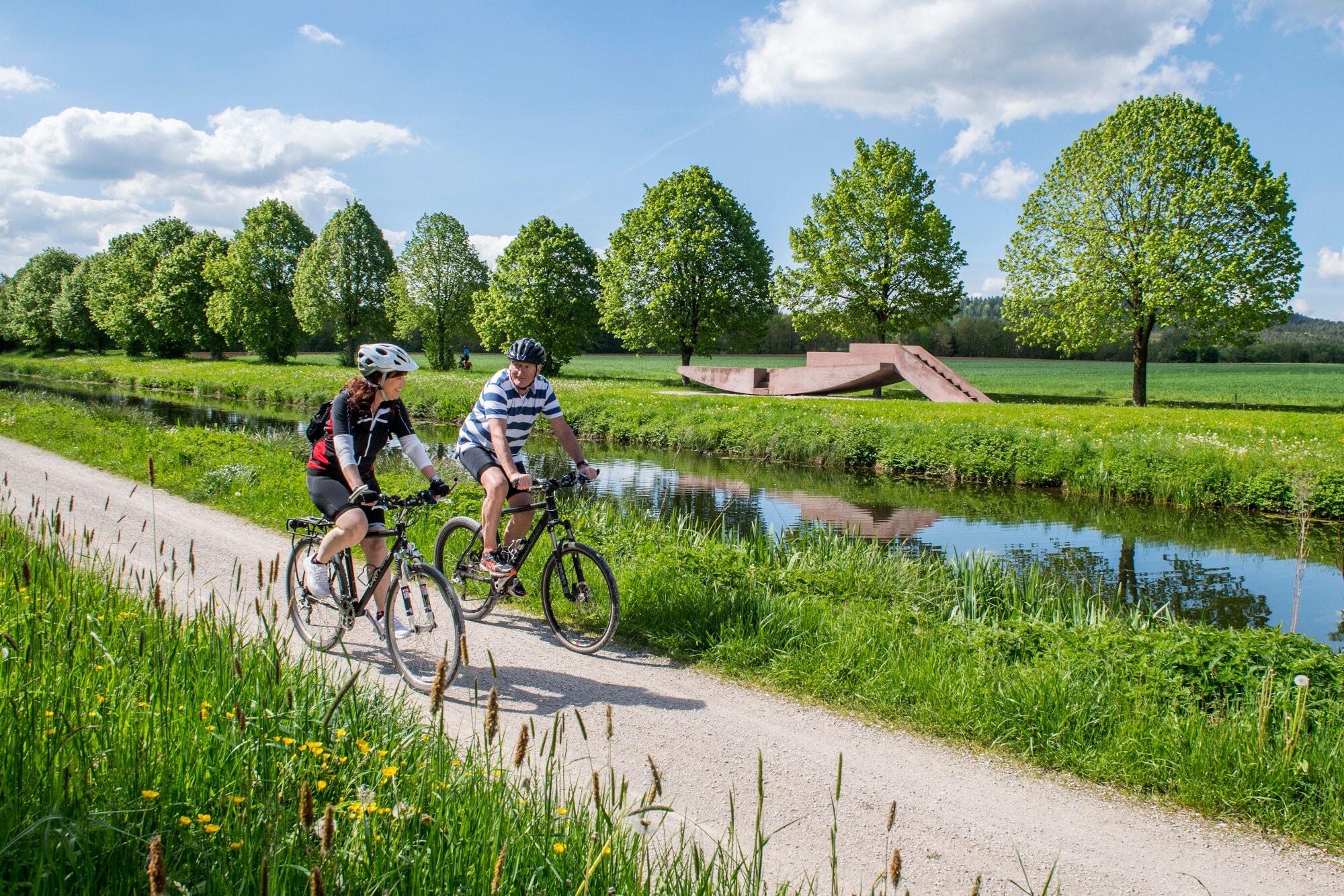 Zwei Personen auf Fahrrädern fahren auf einem Radweg durch eine grüne Landschaft entlang eines Flusses.