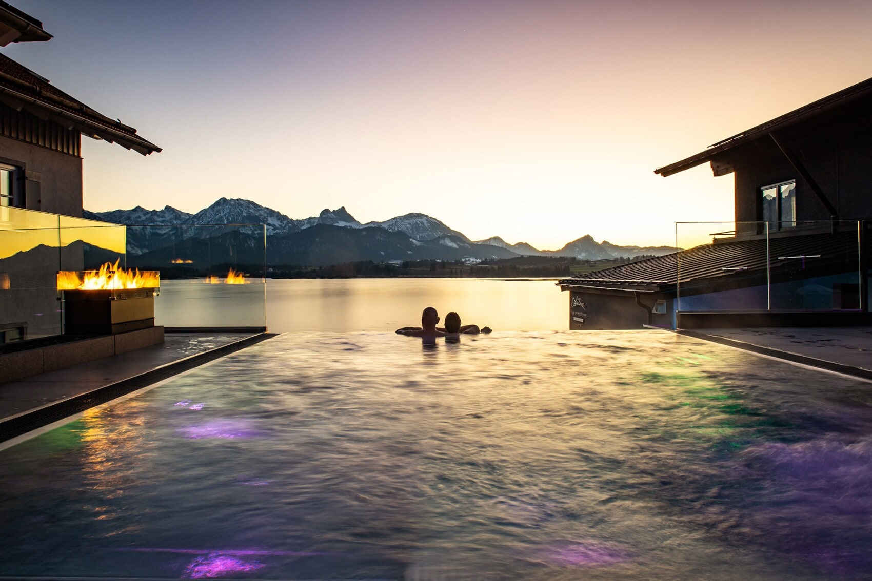 Zwei Personen in einem Infinity-Pool schauen über einen See in eine Berglandschaft bei Sonnenuntergang Zwei Personen in einem Infinity-Pool schauen über einen See in eine Berglandschaft bei Sonnenuntergang