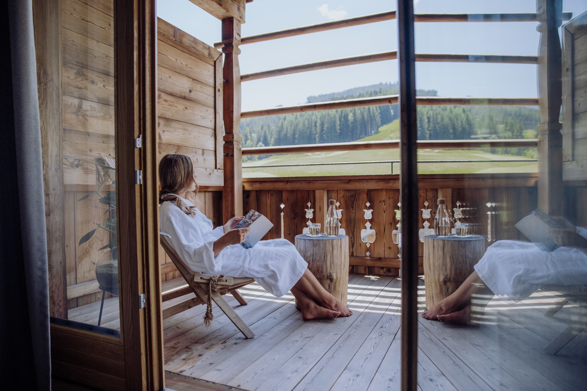 Eine Frau in weißem Bademantel sitzt mit einem Buch auf einer Terrasse eines Holzchalets mit Blick in die Berge Eine Frau in weißem Bademantel sitzt mit einem Buch auf einer Terrasse eines Holzchalets mit Blick in die Berge
