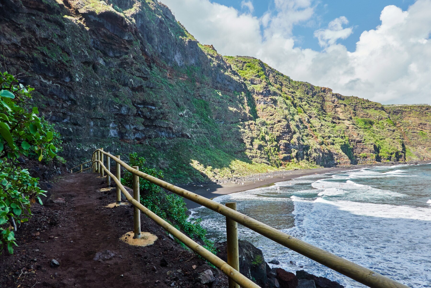 Ein schmaler Wanderweg, der entlang einer grün bewucherten Felsklippe am Meer zu einem kleinen Sandstrand führt