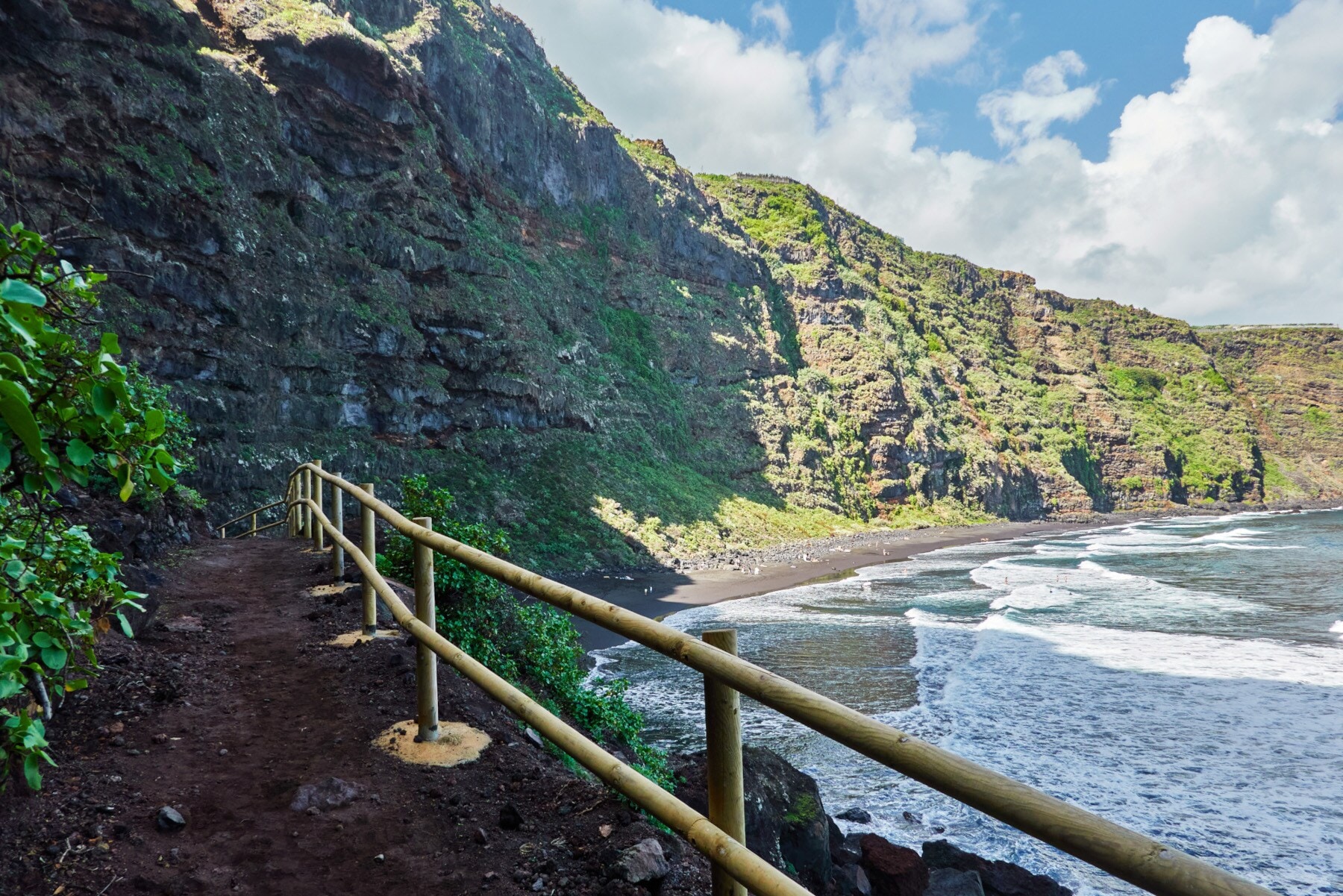 Ein schmaler Wanderweg, der entlang einer grün bewucherten Felsklippe am Meer zu einem kleinen Sandstrand führt Ein schmaler Wanderweg, der entlang einer grün bewucherten Felsklippe am Meer zu einem kleinen Sandstrand führt