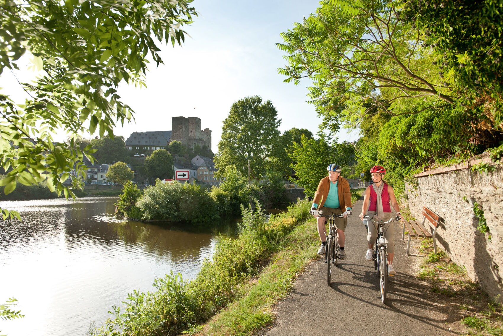 Ein älteres Paar fährt auf Fahrrädern auf einem asphaltierten Radweg entlang eines Flusses, im Hintergrund eine mittelalterliche Burgruine in einer Ortschaft Ein älteres Paar fährt auf Fahrrädern auf einem asphaltierten Radweg entlang eines Flusses, im Hintergrund eine mittelalterliche Burgruine in einer Ortschaft