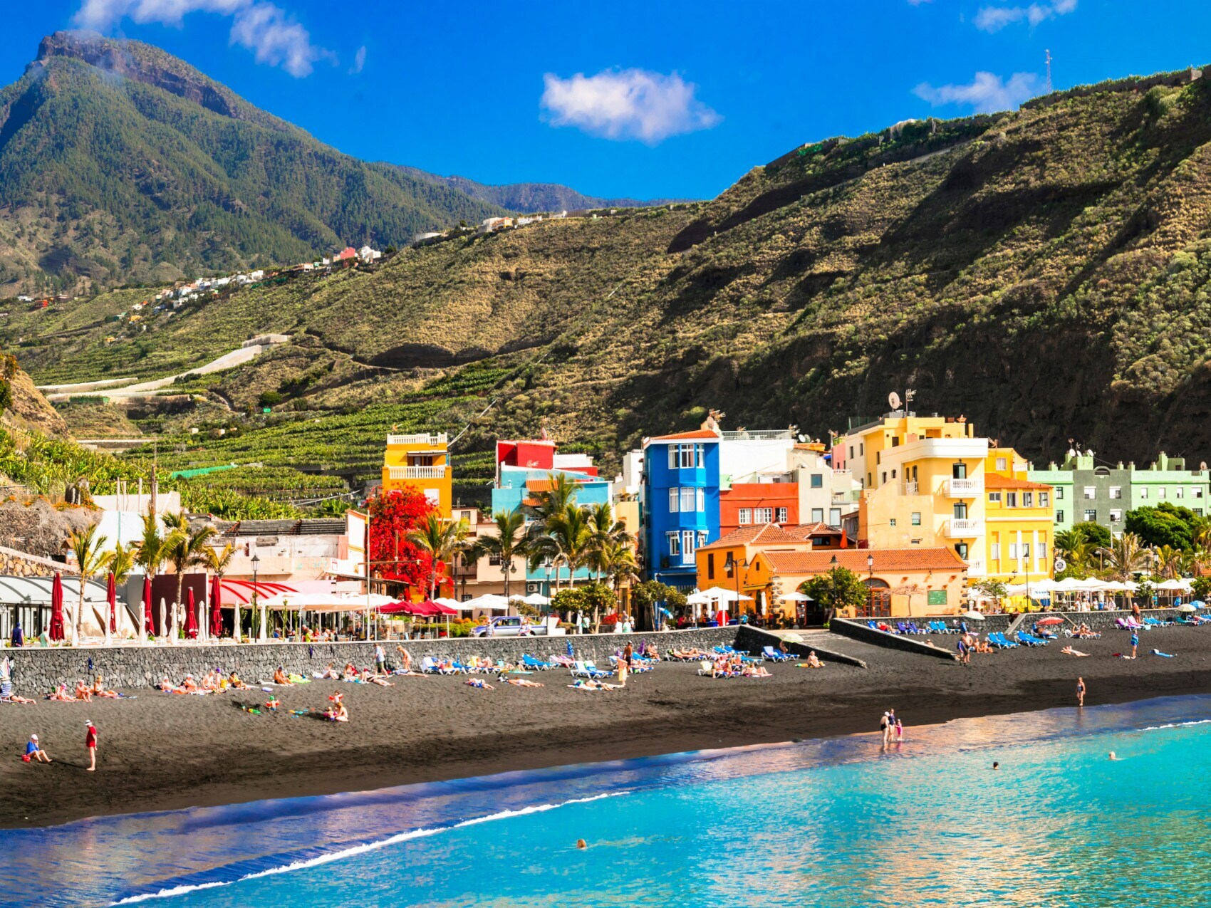 Schwarzer Sandstrand mit Badenden in einem Ort mit bunten Häusern vor Bergpanorama Schwarzer Sandstrand mit Badenden in einem Ort mit bunten Häusern vor Bergpanorama