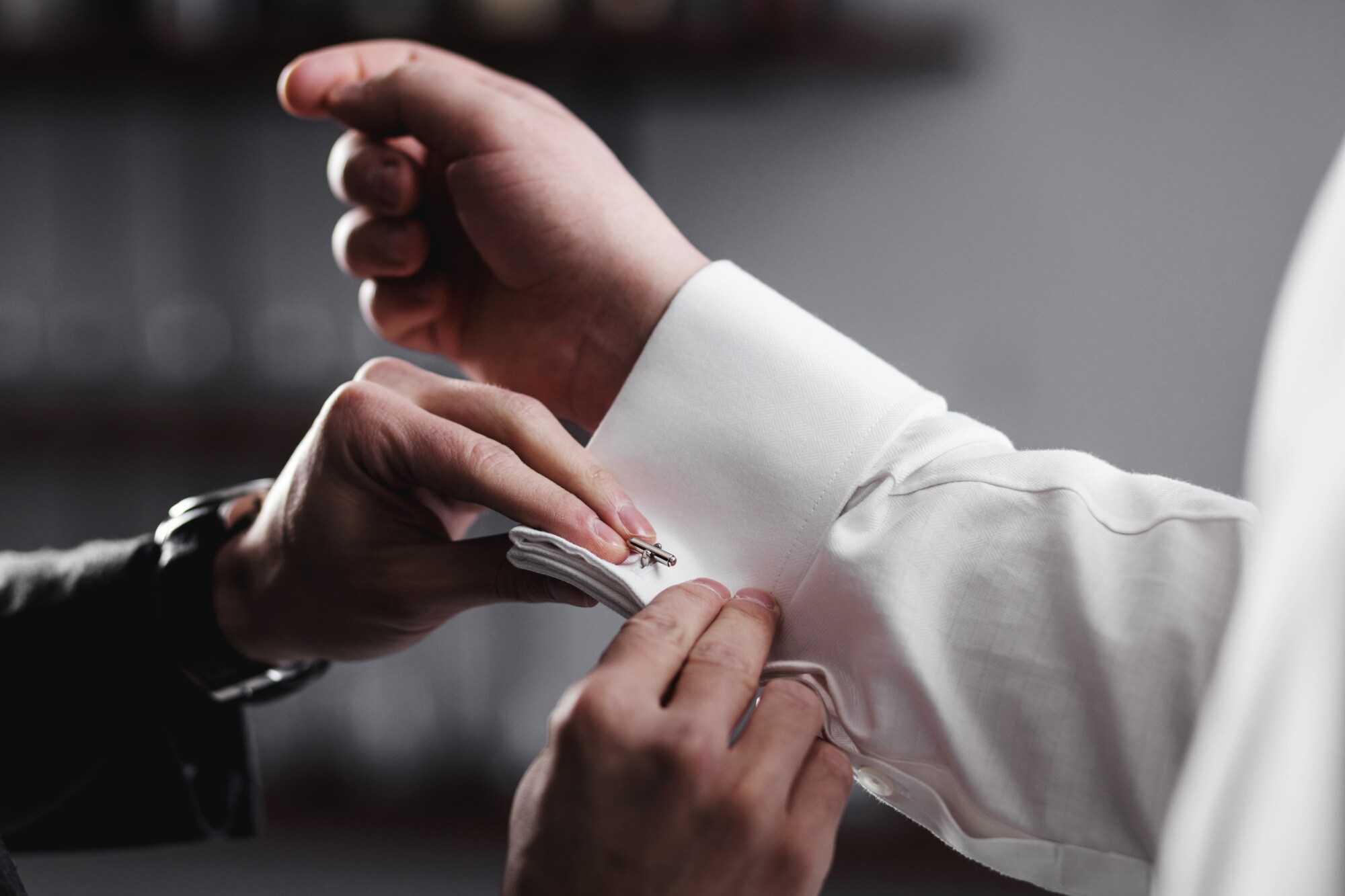 Nahaufnahme von zwei Händen, die einen Manschettenknopf am Hemd einer anderen Person anbringen.