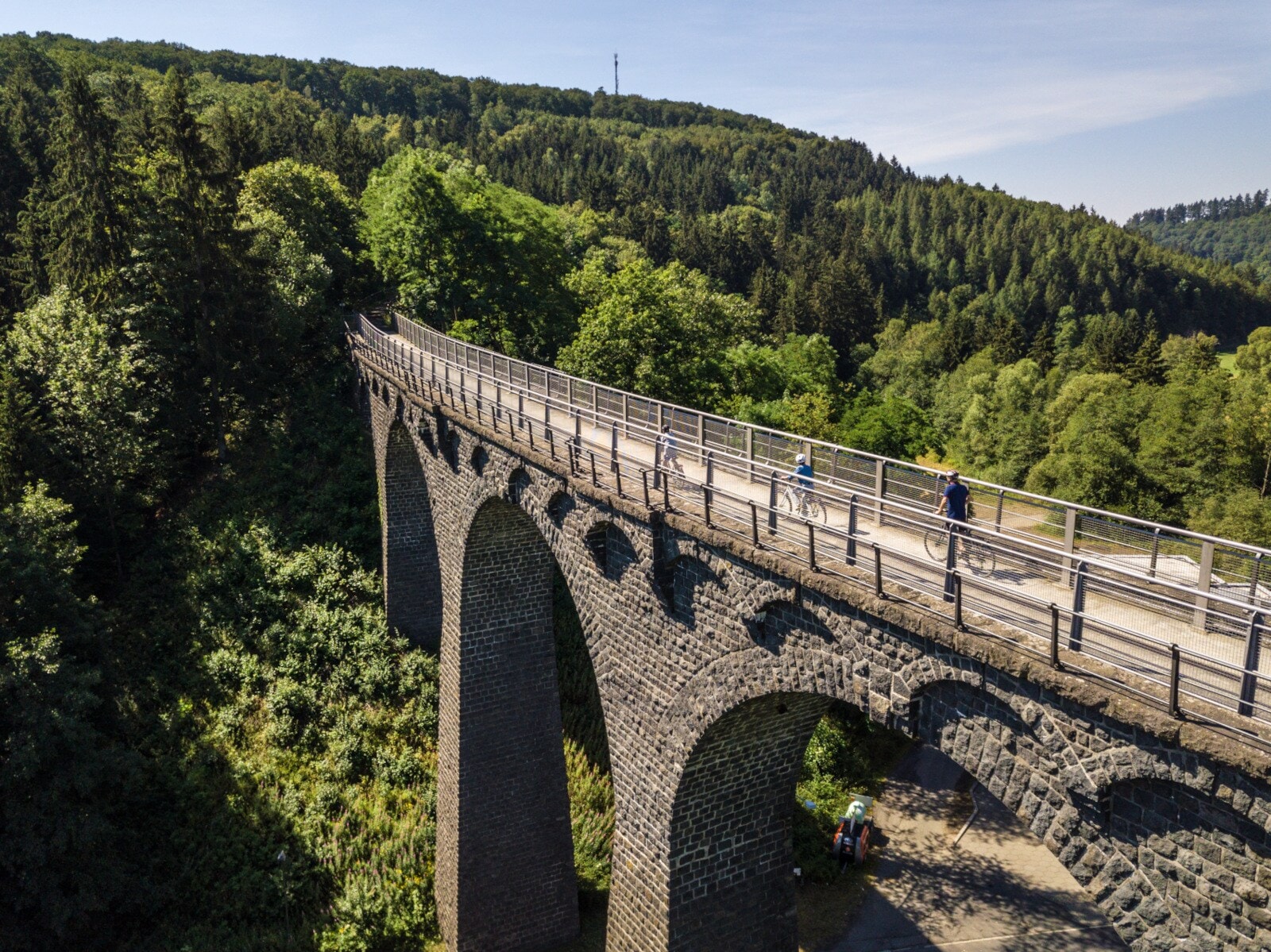 Drei Radfahrende fahren auf einem schmalen Weg auf einem Viadukt in einer hügeligen Waldlandschaft Drei Radfahrende fahren auf einem schmalen Weg auf einem Viadukt in einer hügeligen Waldlandschaft