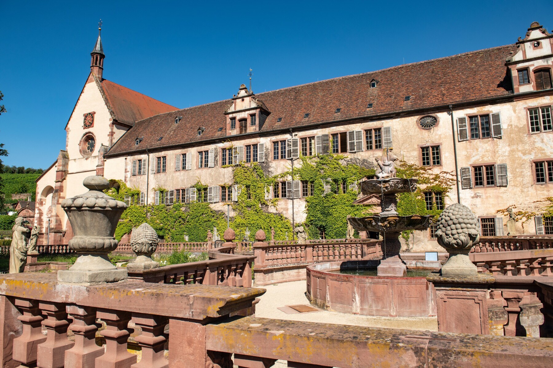 Ein historisches Gebäude, bewachsen mit Grün, davor ein Abteigarten mit steinernen Skulpturen