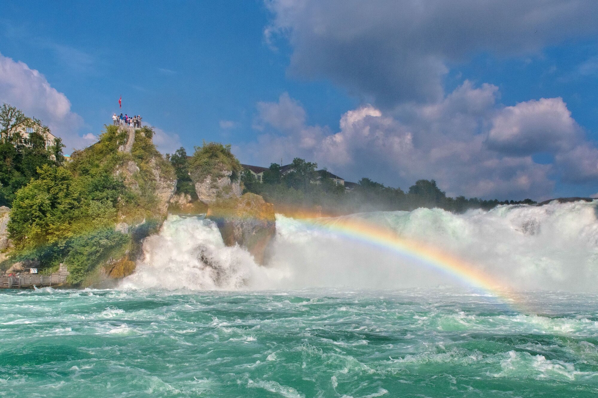 Personen auf einer Aussichtsplattform auf einem Felsen oberhalb eines Wasserfalls, vor dem sich ein Regenbogen gebildet hat