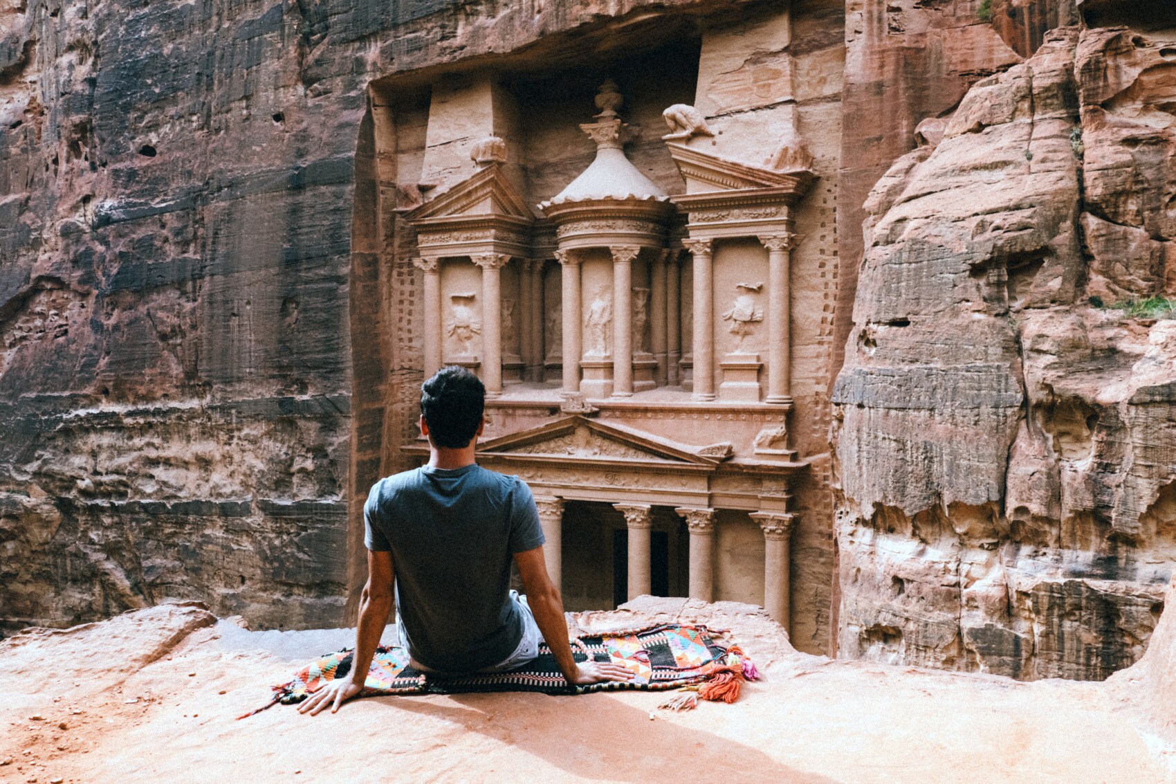 Rückansicht eines auf einem Felsvorsprung sitzenden Mannes vor dem Eingang eines monumentalen Grabtempels in der Felsenstadt Petra Rückansicht eines auf einem Felsvorsprung sitzenden Mannes vor dem Eingang eines monumentalen Grabtempels in der Felsenstadt Petra