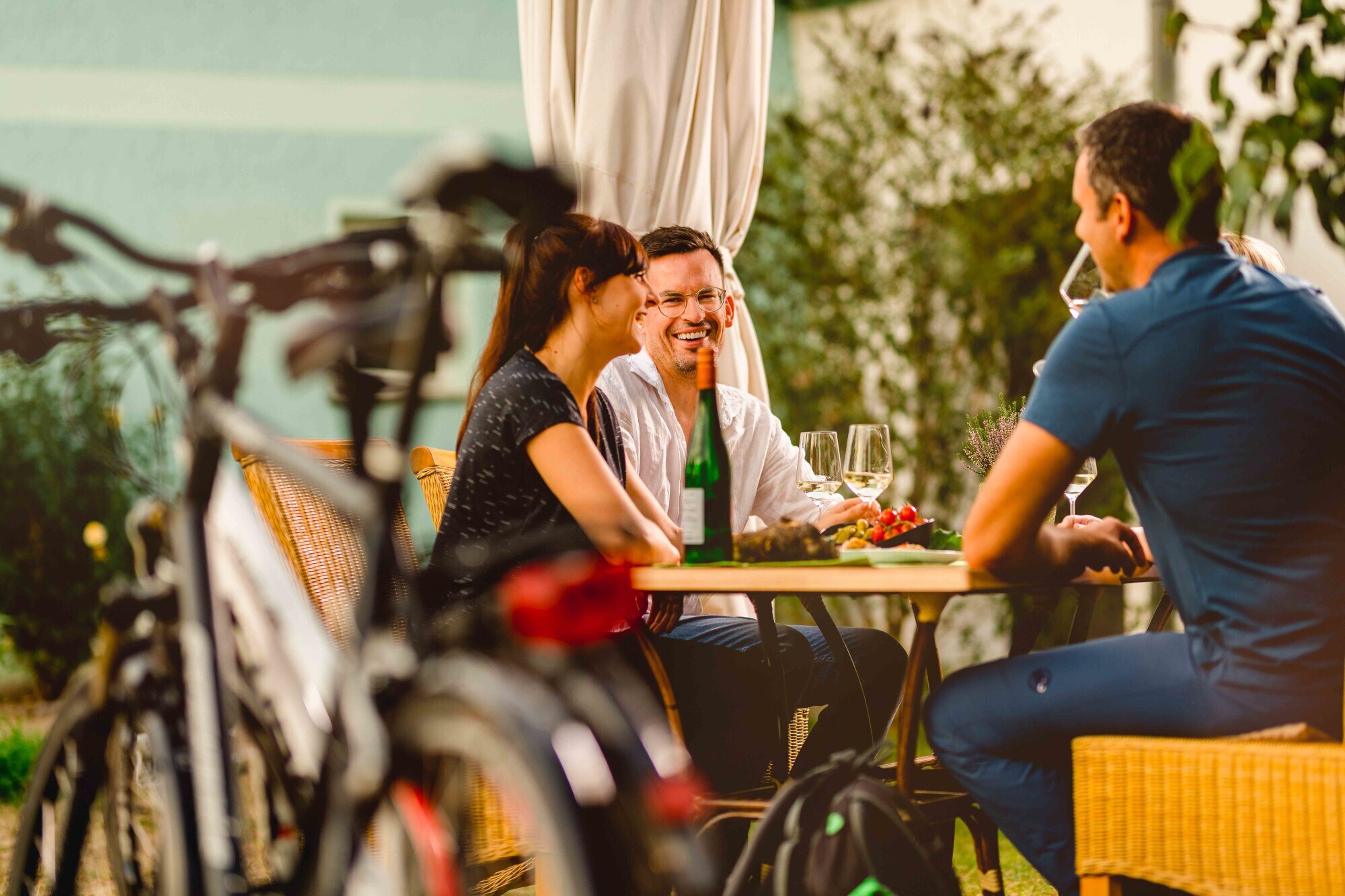 Drei Personen an einem Tisch bei einer Weinverkostung, im Vordergrund geparkte Fahrräder