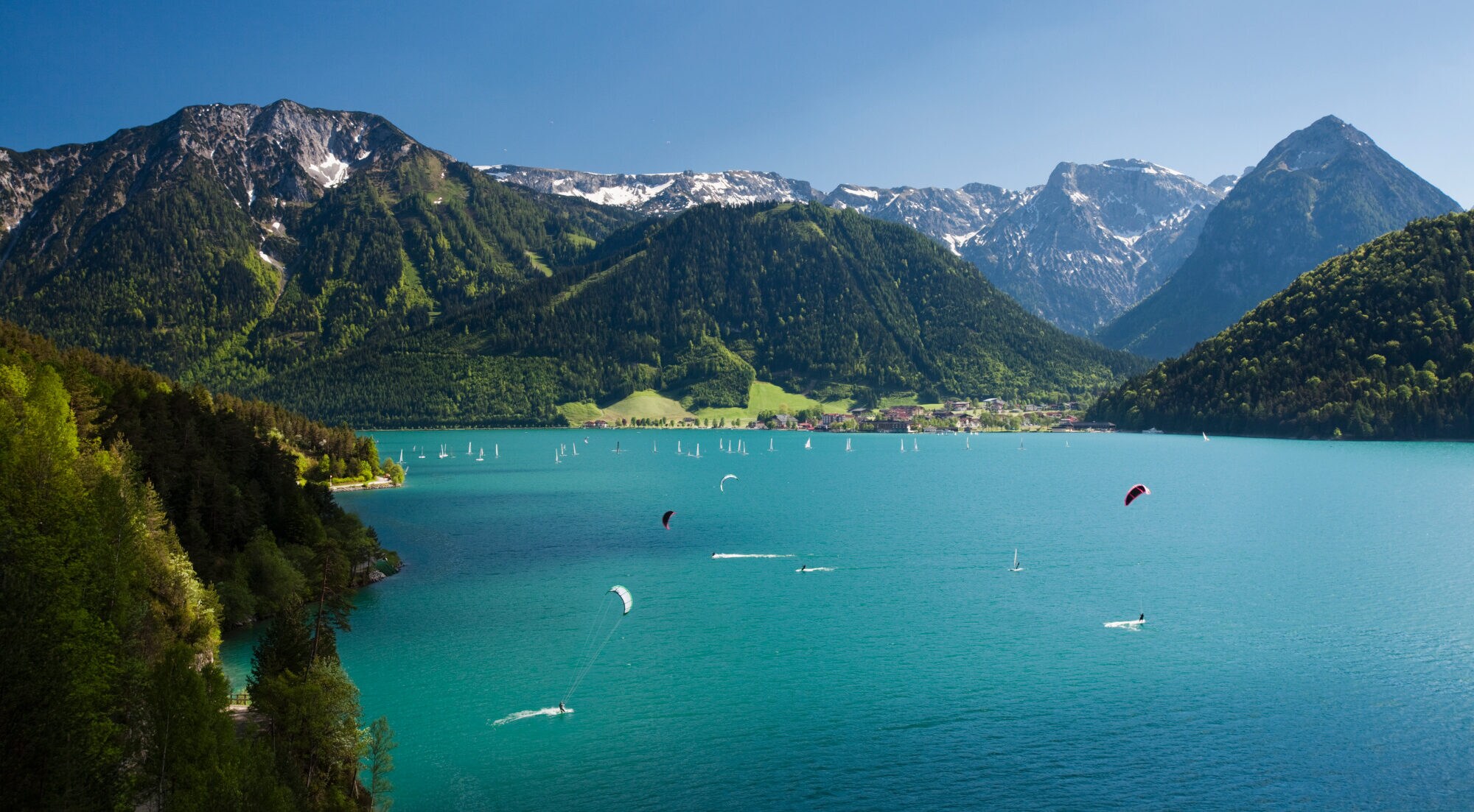 Ein großer See zwischen Bergen, auf dem Wasser zahlreiche Personen, die kitesurfen