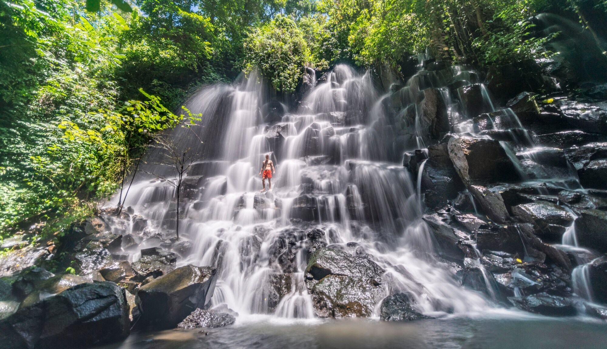Eine Person in roter Badehose steht in einem gestuften Wasserfall Eine Person in roter Badehose steht in einem gestuften Wasserfall