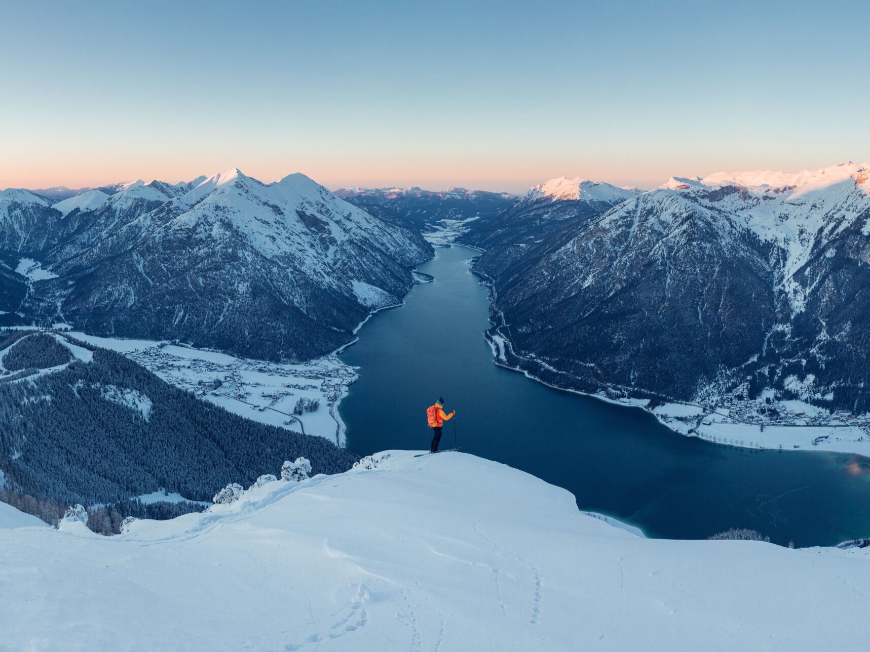Eine Person steht auf einer schneebedeckten Fläche auf einem Berg, im Hintergrund liegt ein langgezogener See zwischen zwei Bergketten