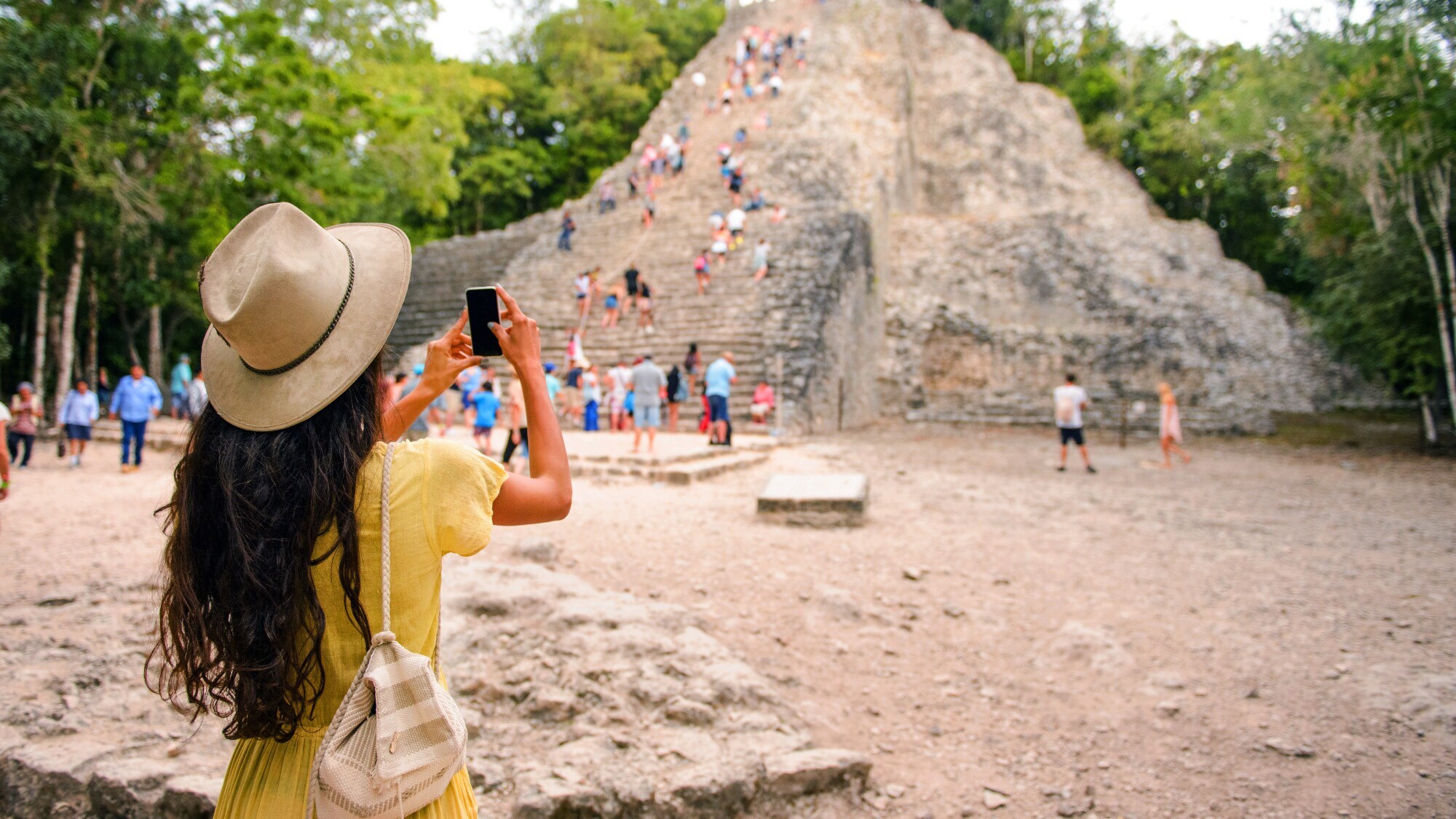 Rückansicht einer Frau mit Strohhut, die mit ihrem Smartphone eine von Personen erklommene Mayapyramide fotografiert