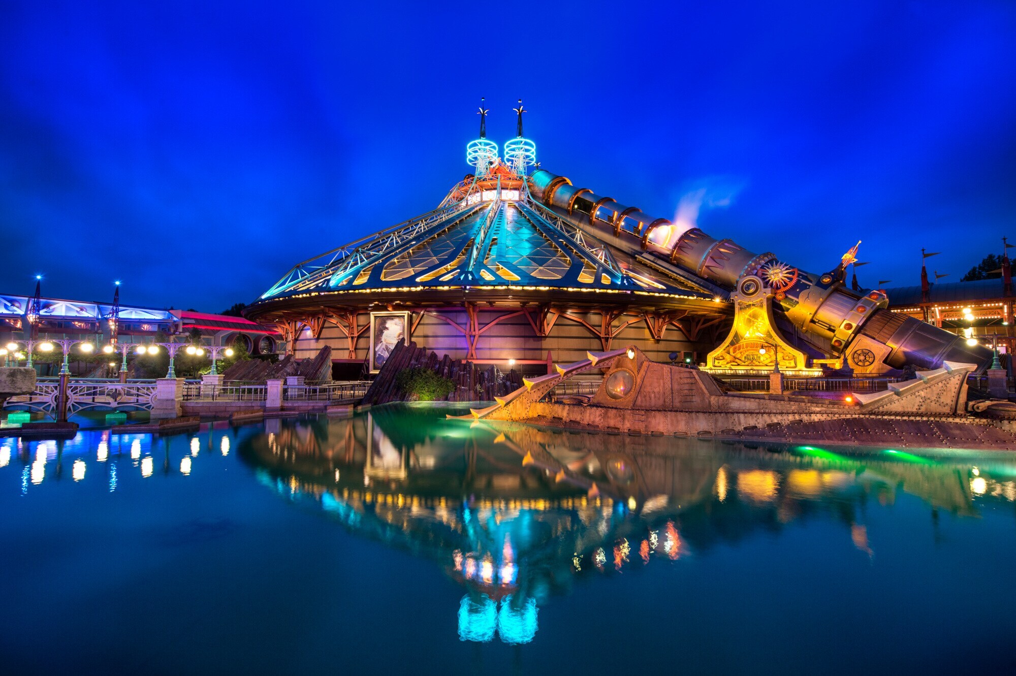 Außenansicht einer beleuchteten Attraktion in Form einer Raumstation an einem See in einem Themenpark Außenansicht einer beleuchteten Attraktion in Form einer Raumstation an einem See in einem Themenpark