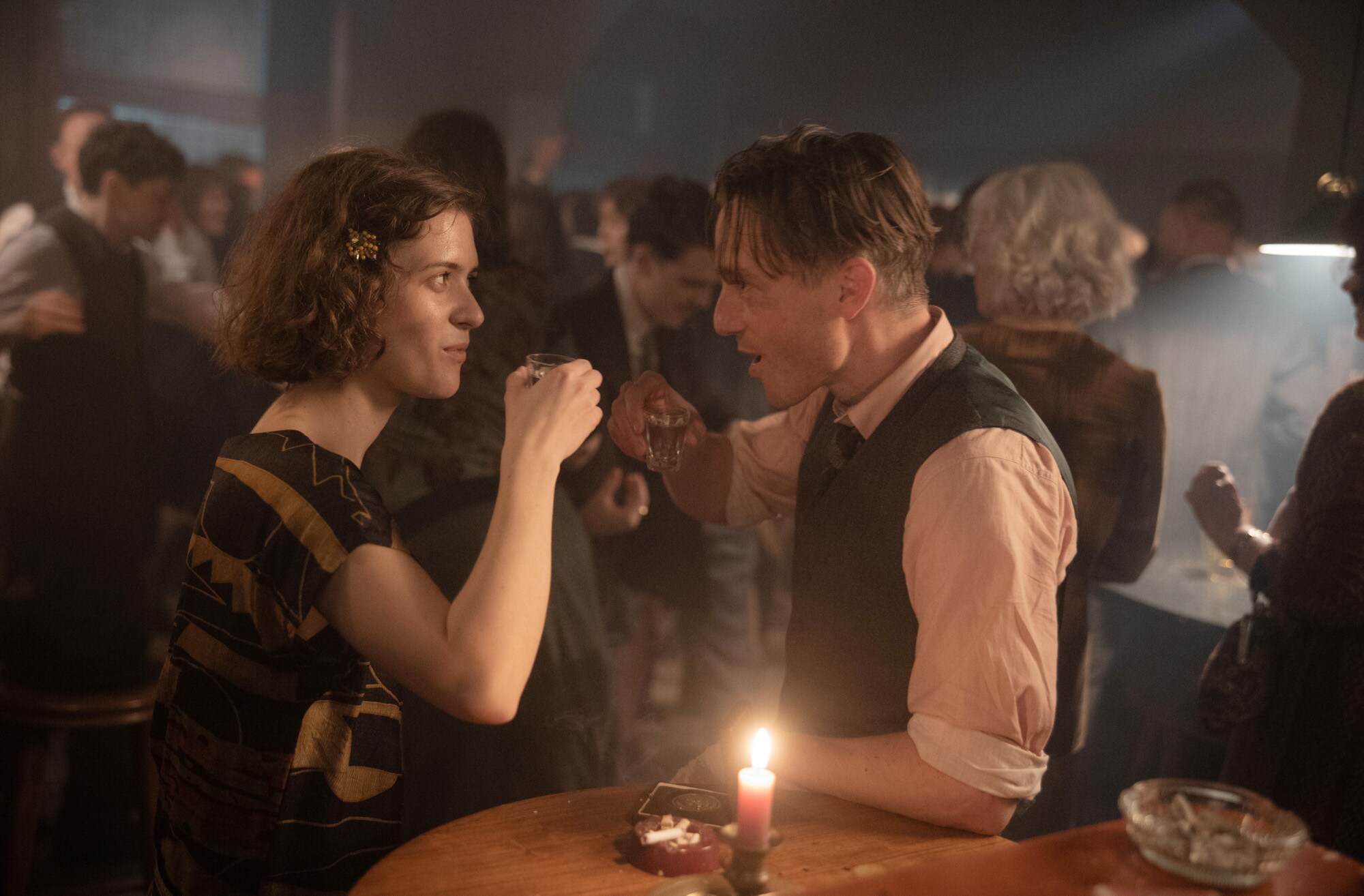 Ein belebter Nachtklub mit Tanzfläche, im Vordergrund ein Mann und eine Frau, die zusammen einen Schnaps trinken