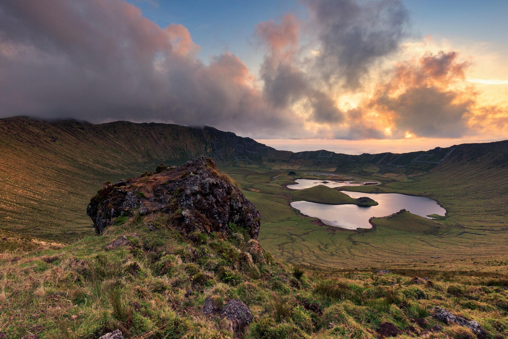 Ein grün bewachsener Vulkankrater mit einer Seenplatte, im Vordergrund ein einzelner großer Felsen Ein grün bewachsener Vulkankrater mit einer Seenplatte, im Vordergrund ein einzelner großer Felsen