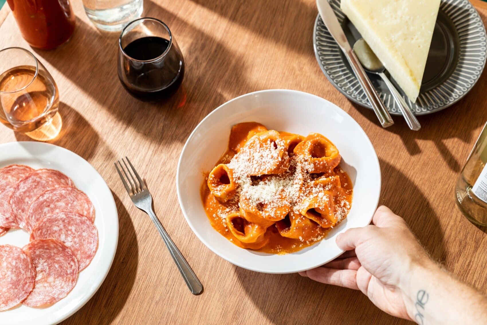 Eine Hand an einem Teller mit Ravioli auf einem eingedeckten Tisch mit Wein und Antipasti