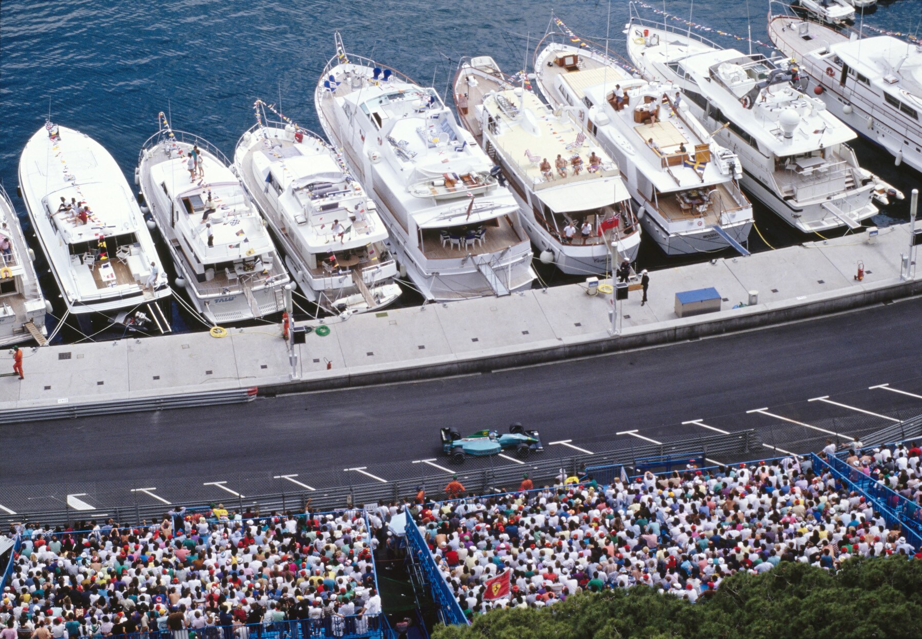 Luftaufnahme eines Rennautos auf einer Rennstrecke zwischen einer Zuschauertribüne und Jachten am Kai