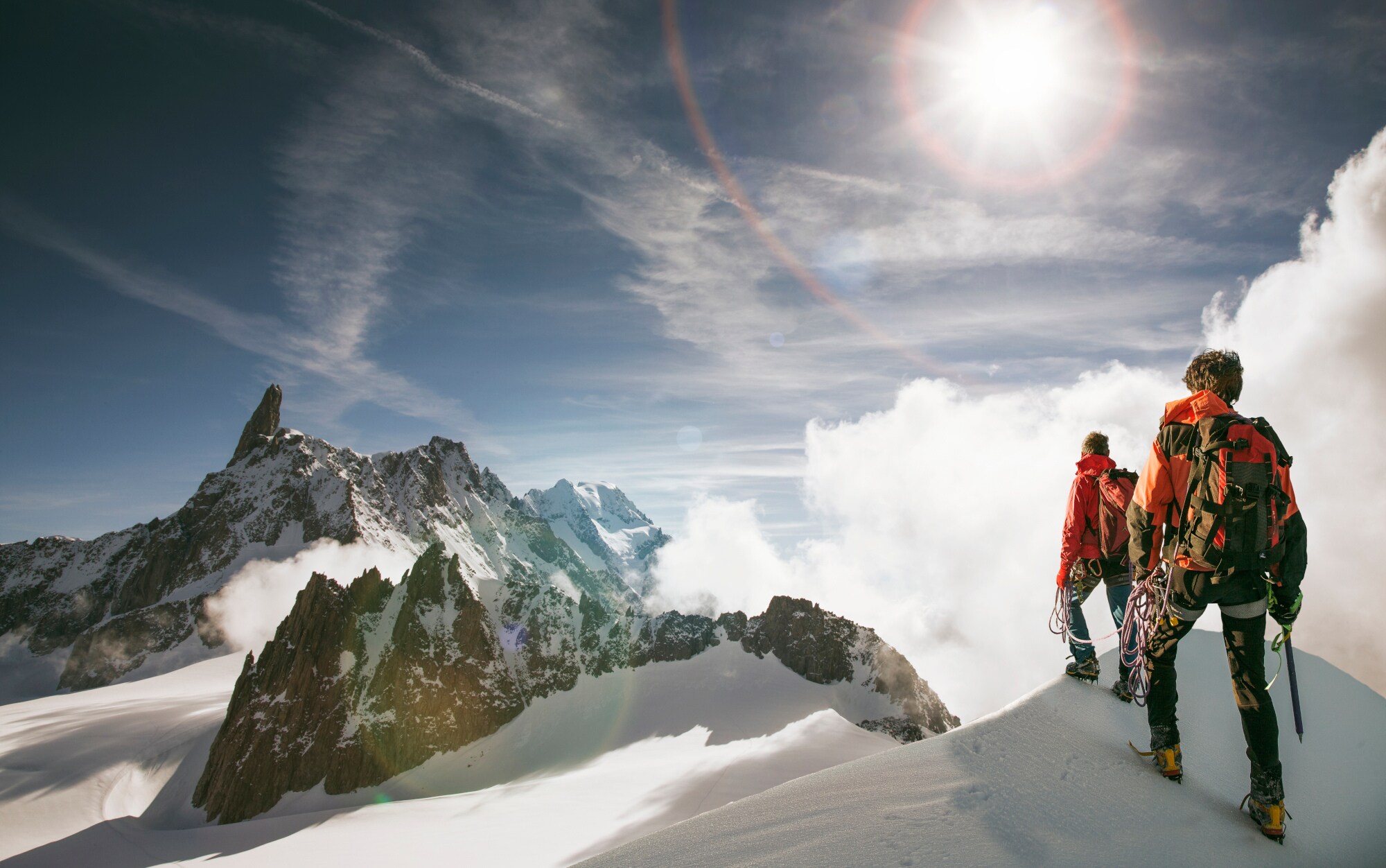 Zwei Personen gehen einen schneebedeckten Bergkamm entlang, im Hintergrund erhebt sich ein noch höheres Bergpanorama