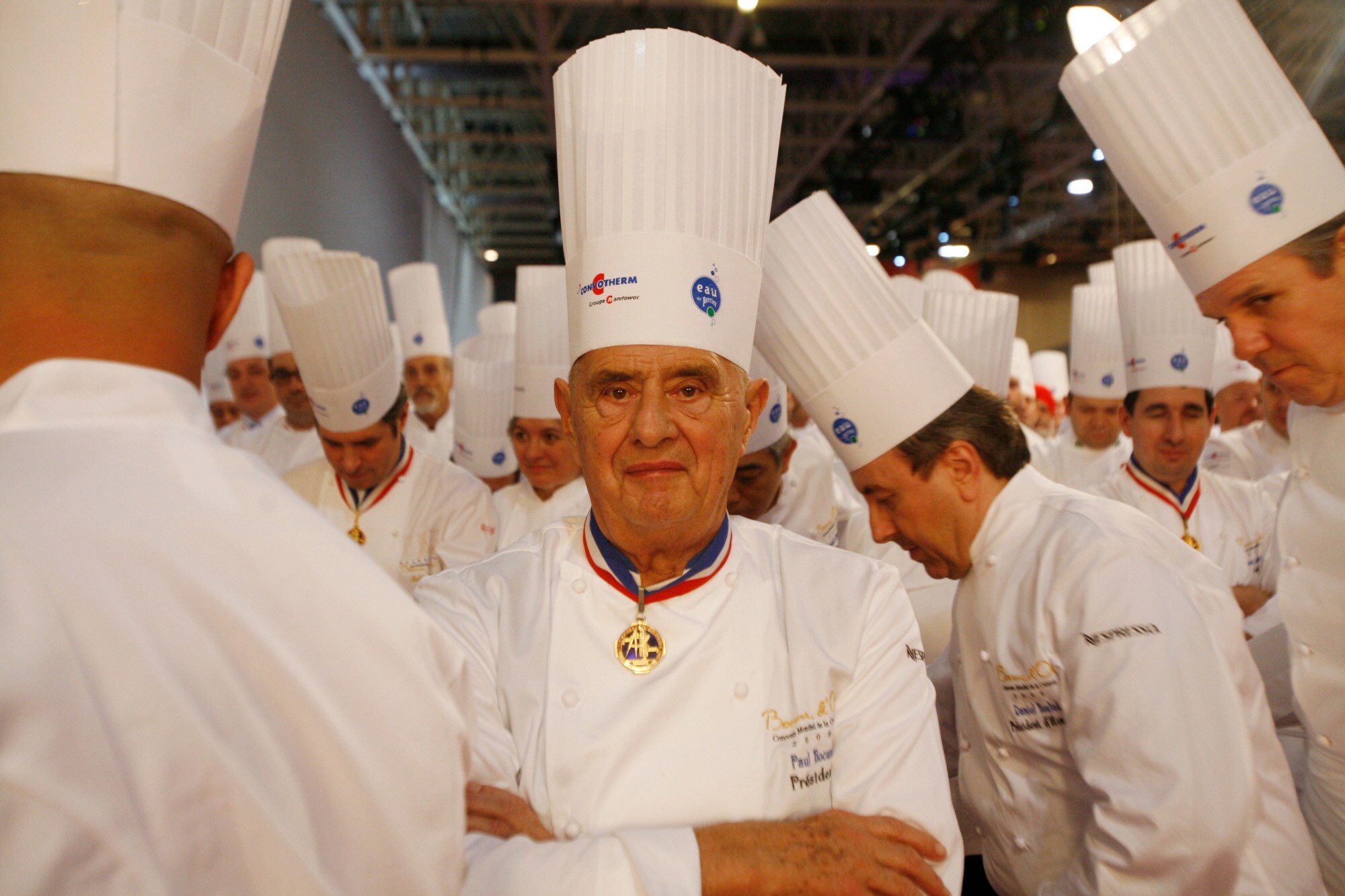 Porträt von Paul Bocuse in Kochuniform inmitten weiterer Köche in einer Halle