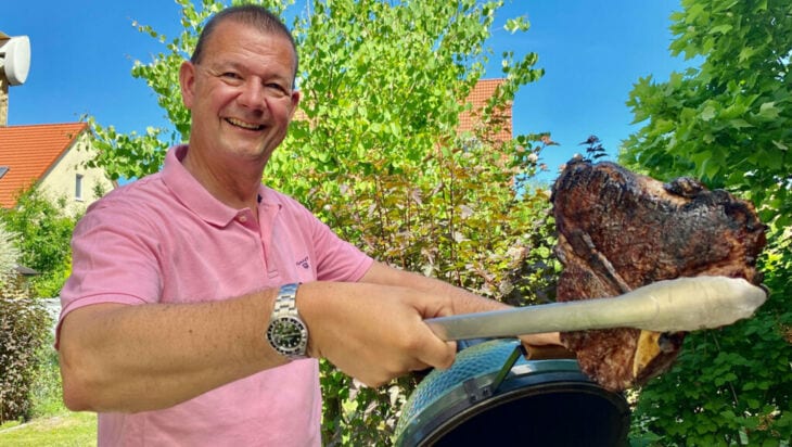 Ein Mann steht an einem Kugelgrill und hält ein fertiges Steak in der Grillzange