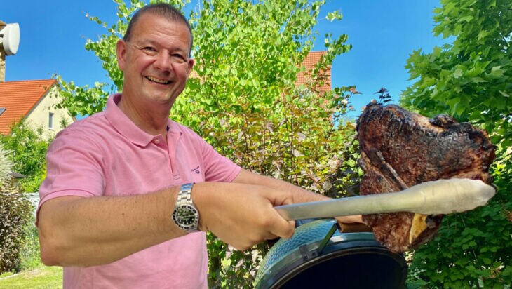 Ein Mann steht an einem Kugelgrill und hält ein fertiges Steak in der Grillzange