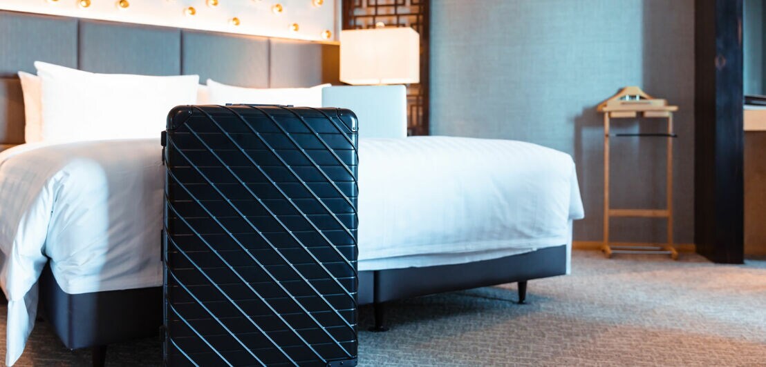 Ein schwarzer Koffer steht in einem Hotelzimmer vor dem Bett