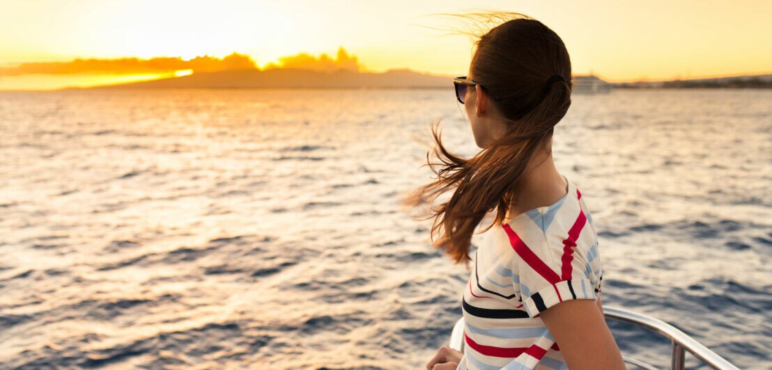 Eine Frau steht auf einem Schiff und blickt in den Sonnenuntergang