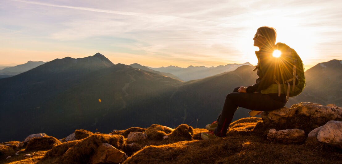Eine Person auf einer felsigen Anhöhe schaut sich den Sonnenuntergang an, im Hintergrund Berggipfel