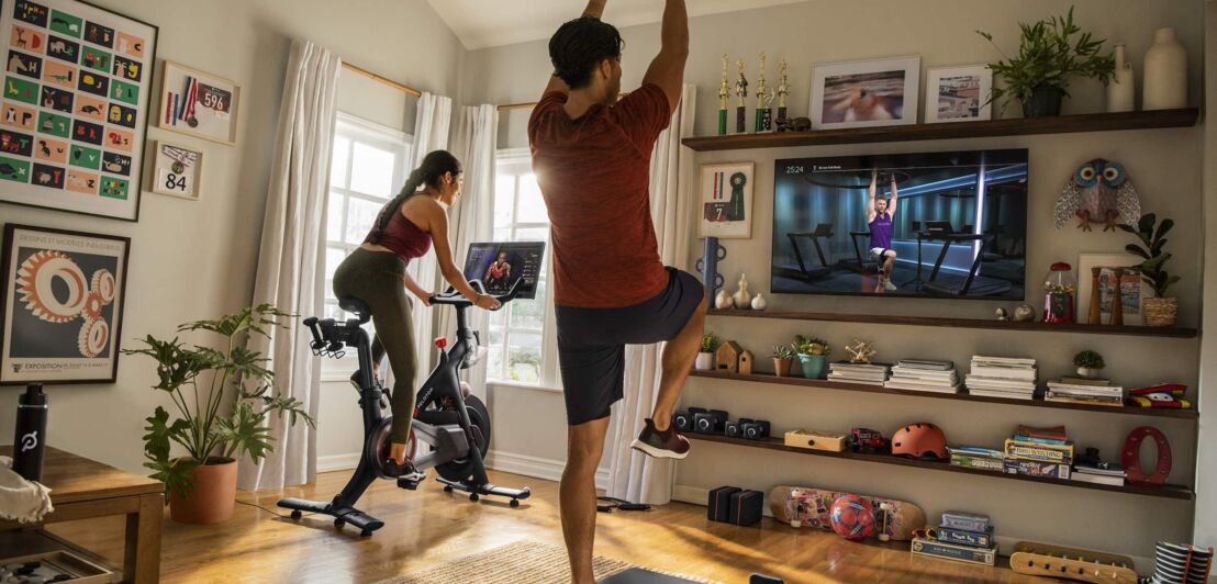 Eine Frau auf einem Heimtrainer-Fahrrad und mit Bildschirm, daneben ein Mann mit einer Hantel in der Hand vor einem Flatscreen, beide machen Sport in einem Wohnzimmer