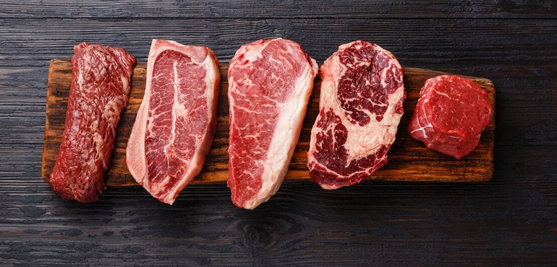 Fünf rohe Steaks auf einem schmalen, langen Holzbrett