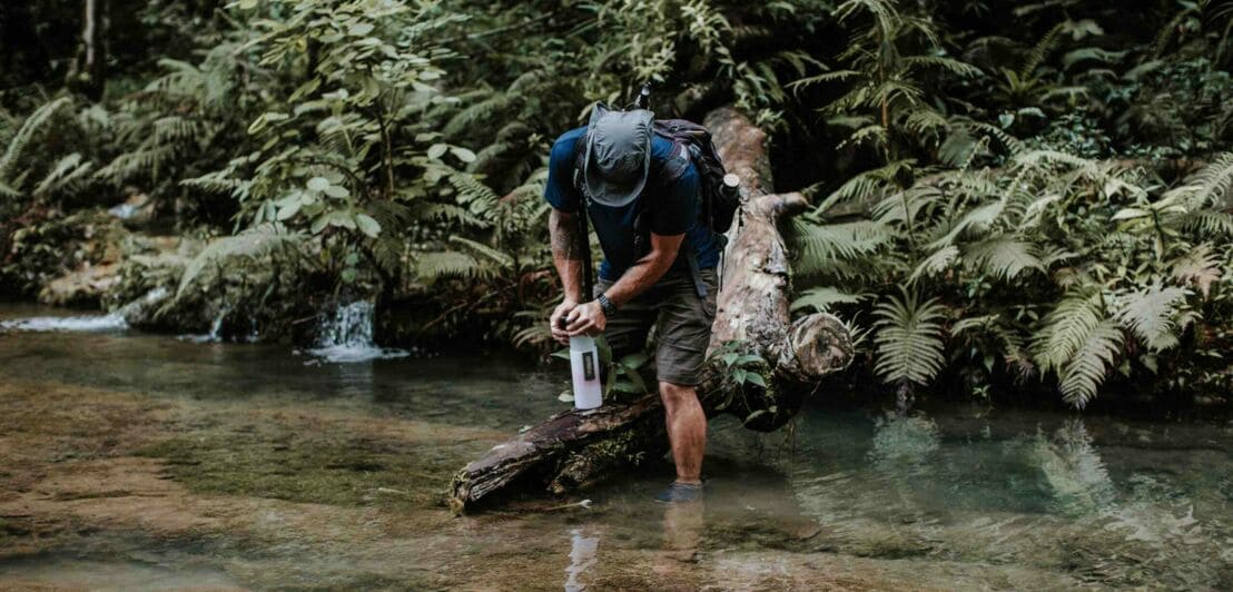 Ein Mann in Shorts steht in einem klaren Fluss und und füllt Wasser in eine Trinkflasche.