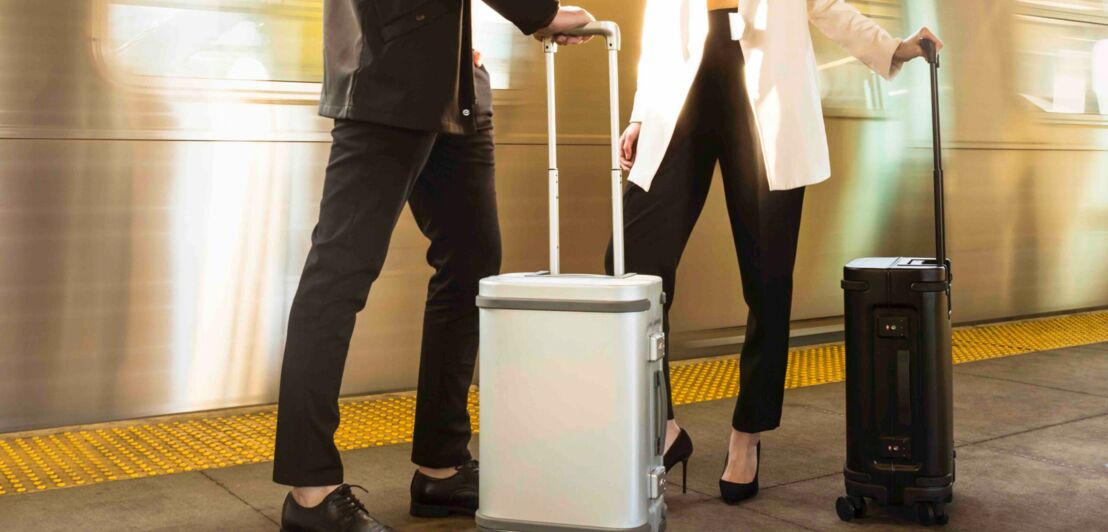 Zwei Menschen mit einem Koffertrolley in der Hand stehen am Gleis einer U-Bahn.