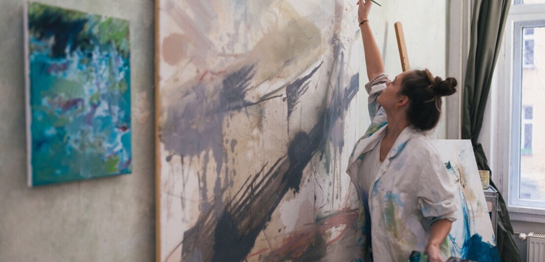 Eine Frau malt mit einem Pinsel auf eine Leinwand die an der Wand hängt