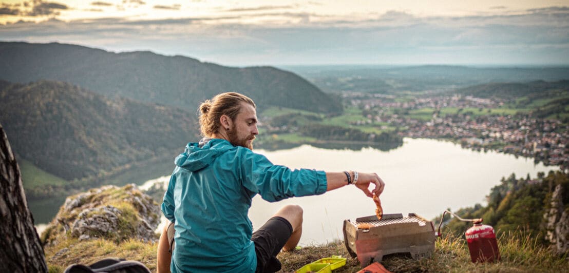 Ein Mann sitzt auf einem Berg mit Blick auf einen See während er ein Stück Fleisch auf den Grill legt