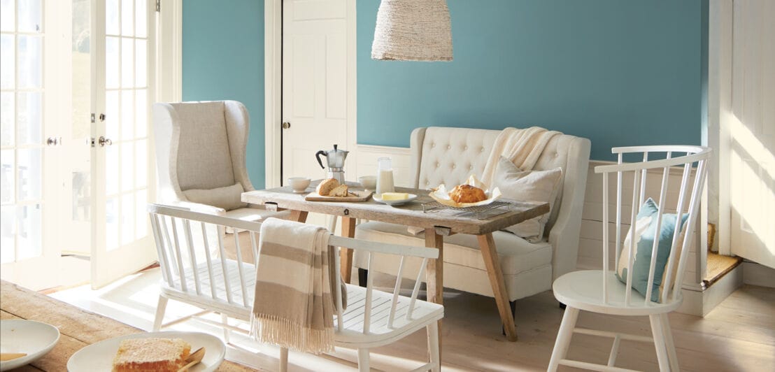 Ein Essbereich mit weißen, hellen Möbeln vor einer hellen, petrolblauen Wand