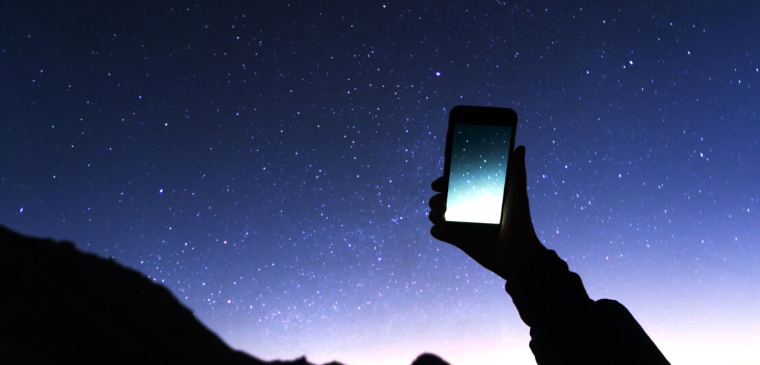 Eine Person hält ein Smartphone mit Sternenfoto vor den Sternenhimmel