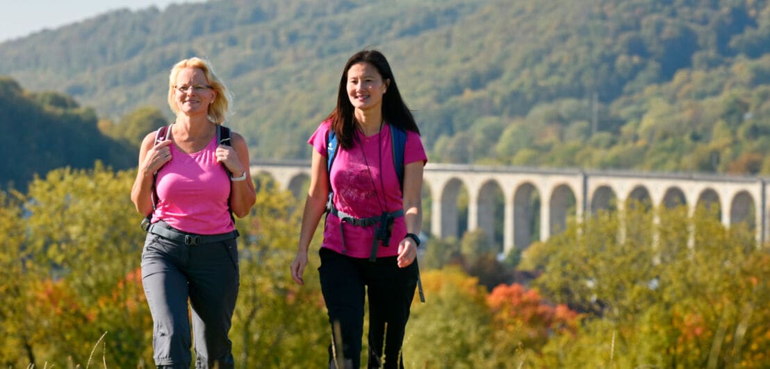 Zwei Frauen wandern durch ein hügelige, waldige Landschaft, im Hintergrund der Viadukt
